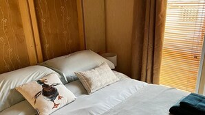 Ein ordentlich gemachtes Bett mit Kissen und gefalteten Handtüchern, ein Nachttisch und ein Fenster mit Jalousien.