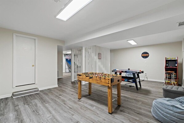 A room with a foosball table, a ping pong table, and a bookshelf.