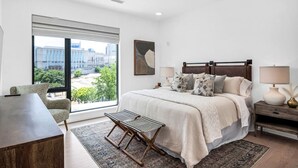 A modern bedroom with a large window, a bed with multiple pillows, a bedside table with a lamp, and a small chair.