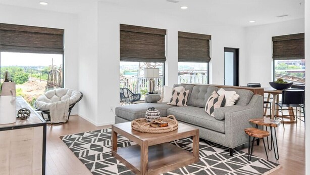 A modern living room with a grey sofa, a white armchair, and a wooden coffee table. There are large windows with dark blinds, a patterned rug, and a view of greenery outside.