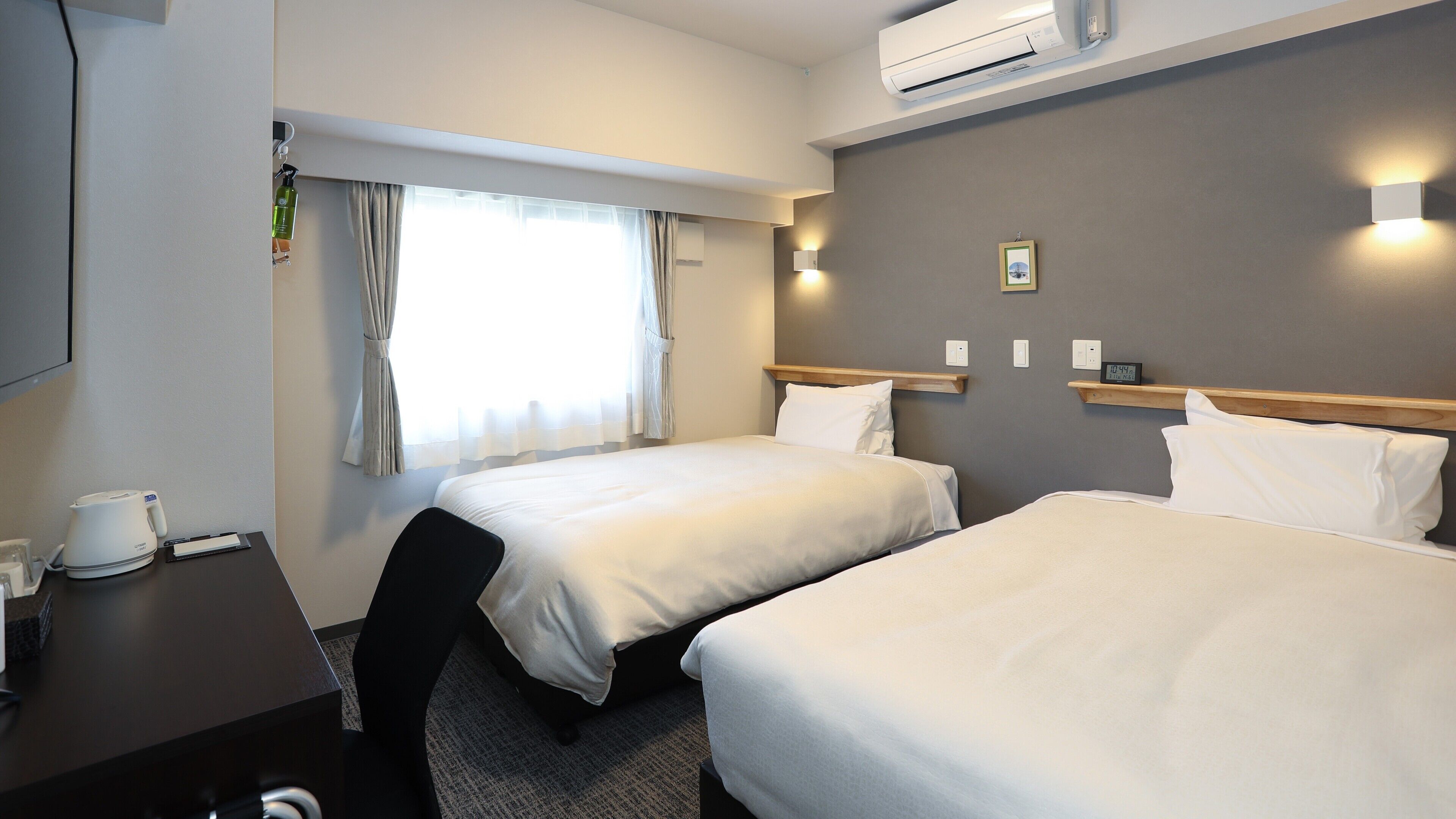 Ein Hotelzimmer mit zwei Betten, einem Schreibtisch mit Wasserkocher, einer Uhr und einem Fenster mit Vorhängen.