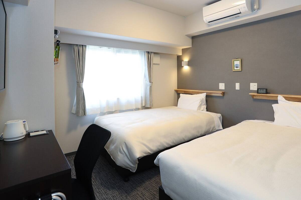 Ein Hotelzimmer mit zwei Betten, einem Schreibtisch mit Wasserkocher, einer Uhr und einem Fenster mit Vorhängen.