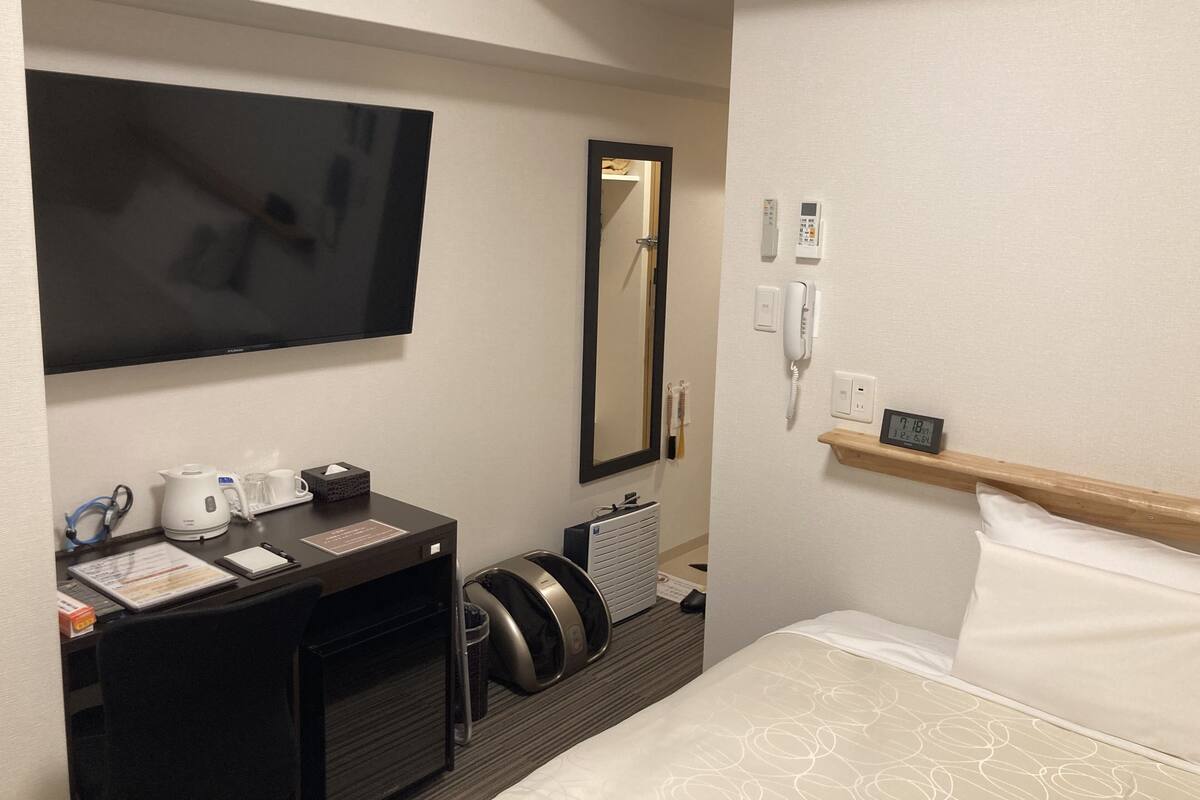 Ein Hotelzimmer mit einem Bett, einem Schreibtisch mit Flachbildfernseher, einem Spiegel, einem Telefon und einem kleinen Regal mit einer Uhr.