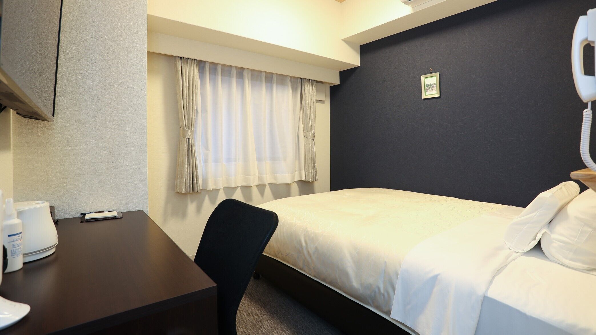 Una habitación de hotel con cama, escritorio, silla, teléfono y ventana con cortinas.