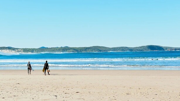 Una playa con dos personas caminando hacia el océano, llevando tablas de surf.