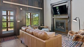 Un salón moderno con un sofá de cuero, chimenea, televisión y ventanas amplias con vistas al exterior.