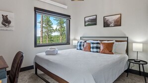 Un dormitorio ordenado con una cama grande, un escritorio y una silla. Hay una ventana con vistas a árboles y montañas.