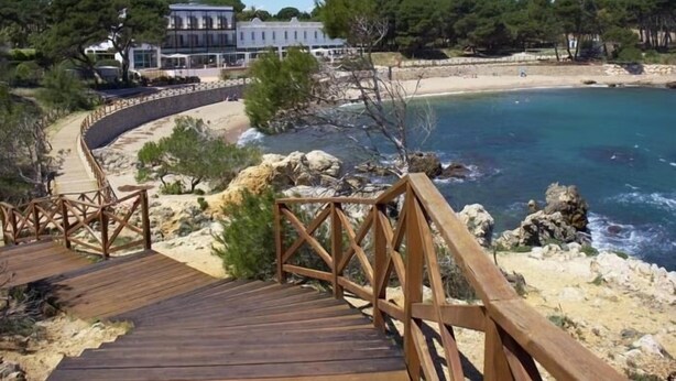 Una pasarela de madera que lleva a una playa con agua azul cristalina, rodeada de terreno rocoso y árboles.