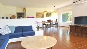 Una sala de estar con un sofá azul, una mesa de centro redonda, un comedor con sillas y un televisor sobre un mueble de madera.