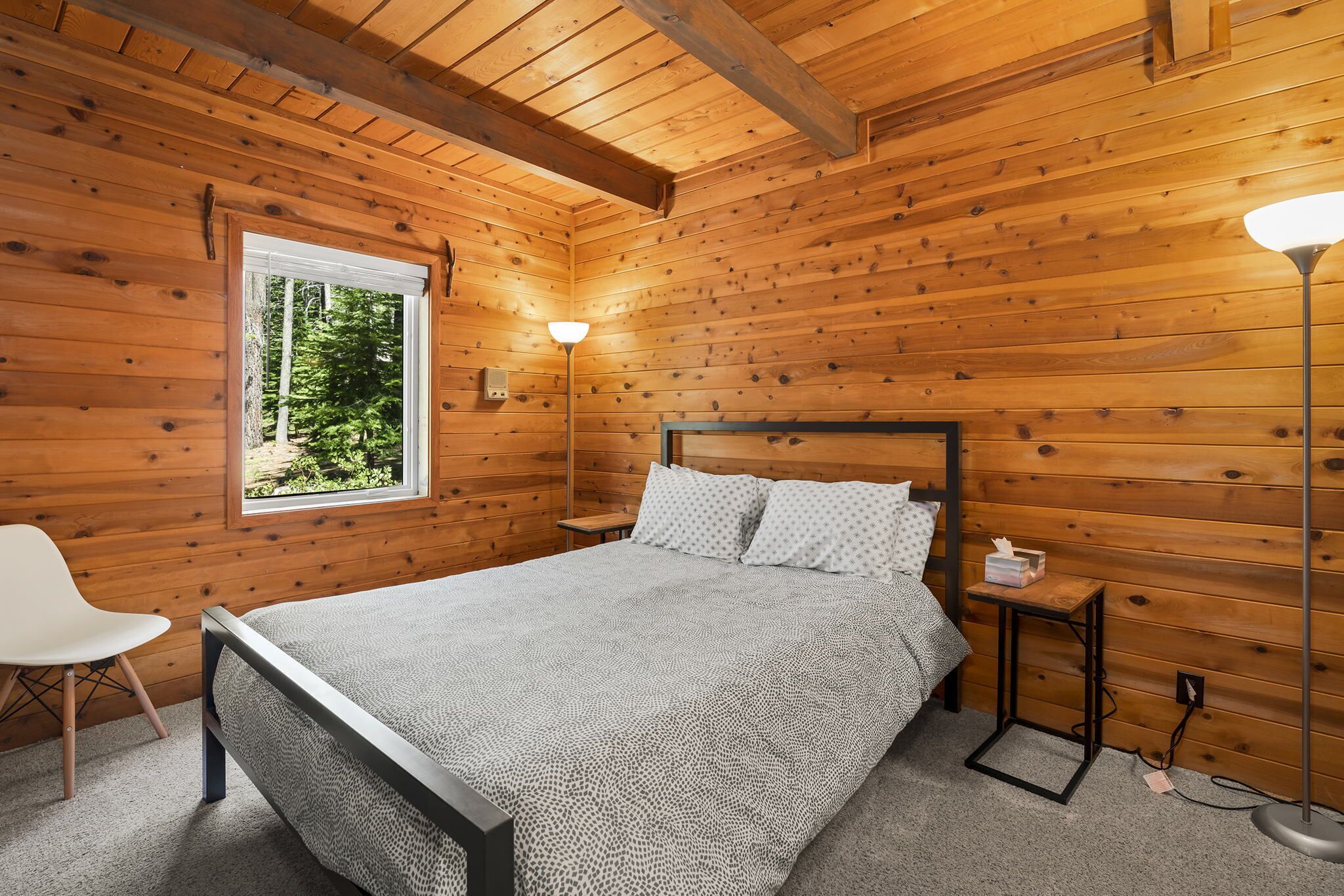 A wooden-paneled bedroom with a bed, bedside table, lamp, and a window with a view of trees.