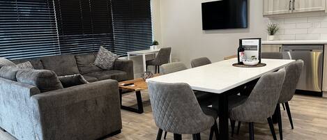 A modern living room with a sofa, chairs, a dining table, and a TV mounted on the wall.