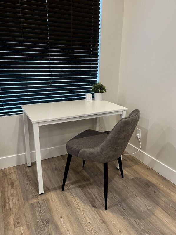 A small, white desk with a grey chair in front of a window with blinds.