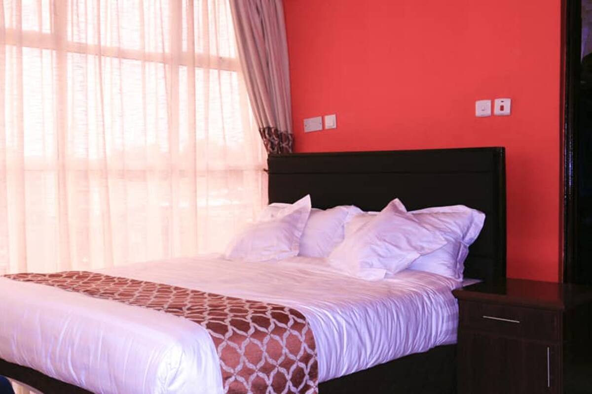 A hotel room with a bed, a nightstand, a red wall, and a window with sheer curtains.
