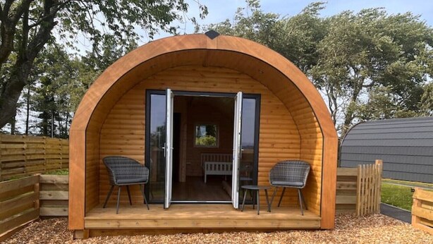 Una estructura abovedada de madera con un puerta de cristal, dos sillas y un banco en el exterior.