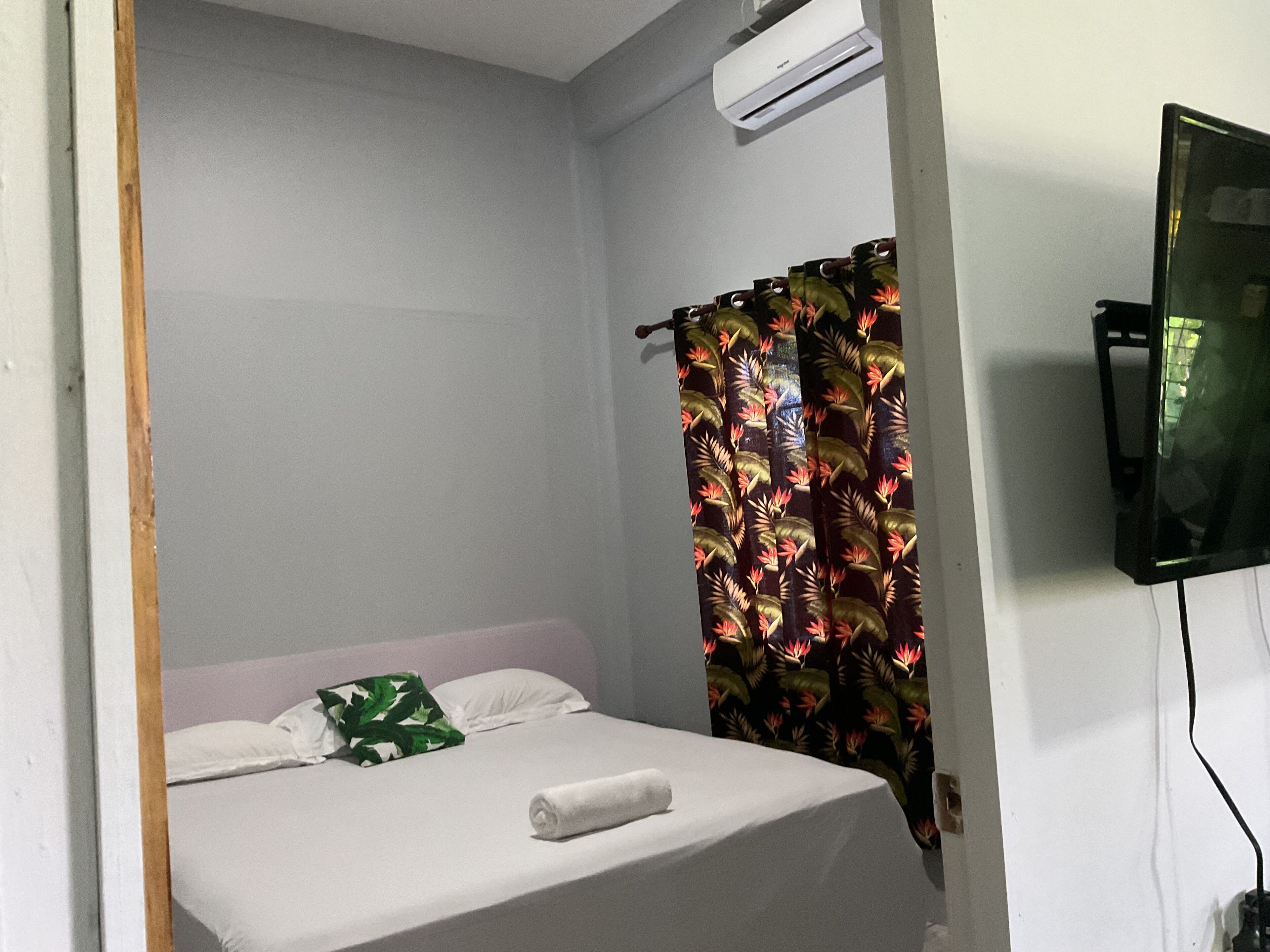 A small, single-bed room with a white bedspread, a green pillow, and a rolled white towel. There is a patterned curtain on the window, and an air conditioning unit mounted on the wall.