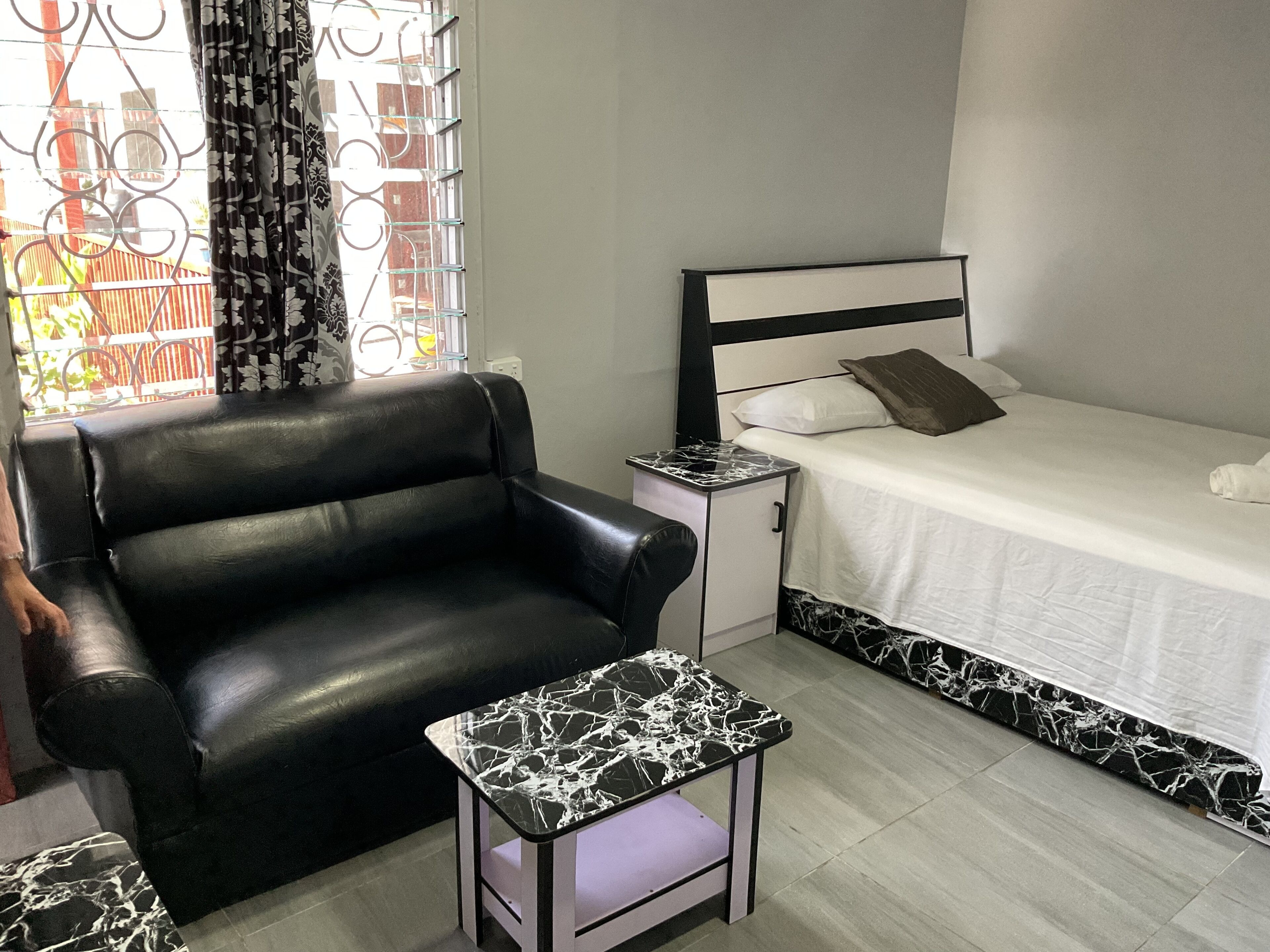 Quarto compacto com sofá de couro preto, uma pequena mesa com padrão marmorizado, cama com roupa de cama branca e janela com cortinas estampadas.