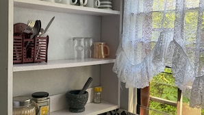 A kitchen with white shelves holding mugs, cups, and a spice rack. A window with lace curtains is visible. - Fox Homestay (Suva)