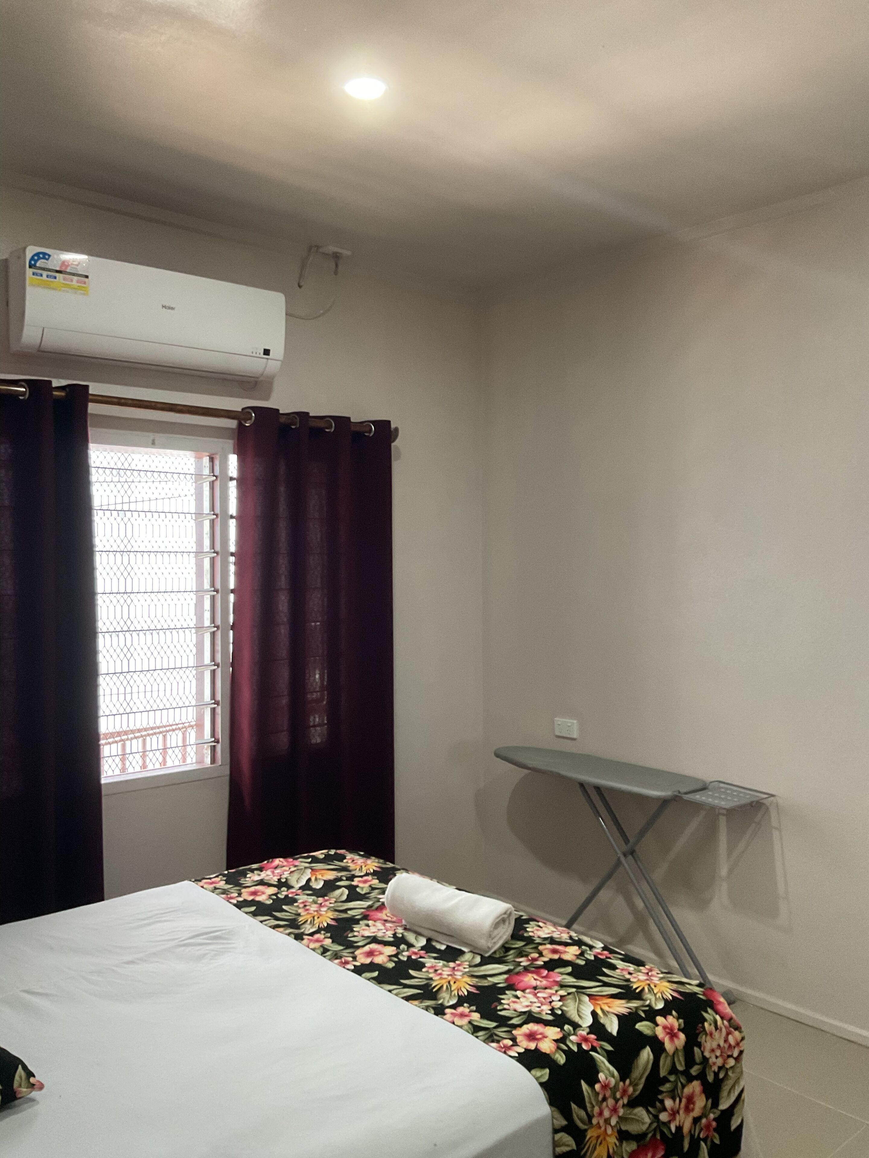A hotel room with a bed, a small table, a window with curtains, and an air conditioning unit.