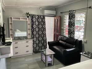 A compact living space with a bed, a white dresser with drawers, a black leather sofa, a small table, and a window with decorative metal grilles.