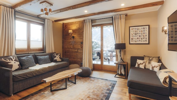 A cozy living room with a sofa, a bench, and a wooden coffee table. There is a large window with curtains, a wall-mounted lamp, and a framed picture on the wall. - Appartement  Streifblick
Wohlfühlappartement für bis zu 6 Personen   (Kitzbühel)