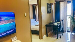 Habitación de hotel con un televisor mostrando una vista panorámica, un comedor con una mesa dispuesta para dos personas y una cama con cortinas.