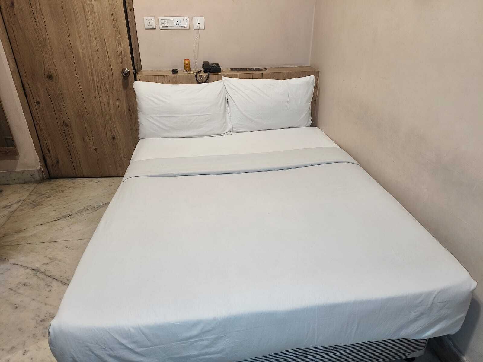 A single bed with a white mattress and pillows, a wooden door to the left, and a wall with electrical outlets and a telephone.