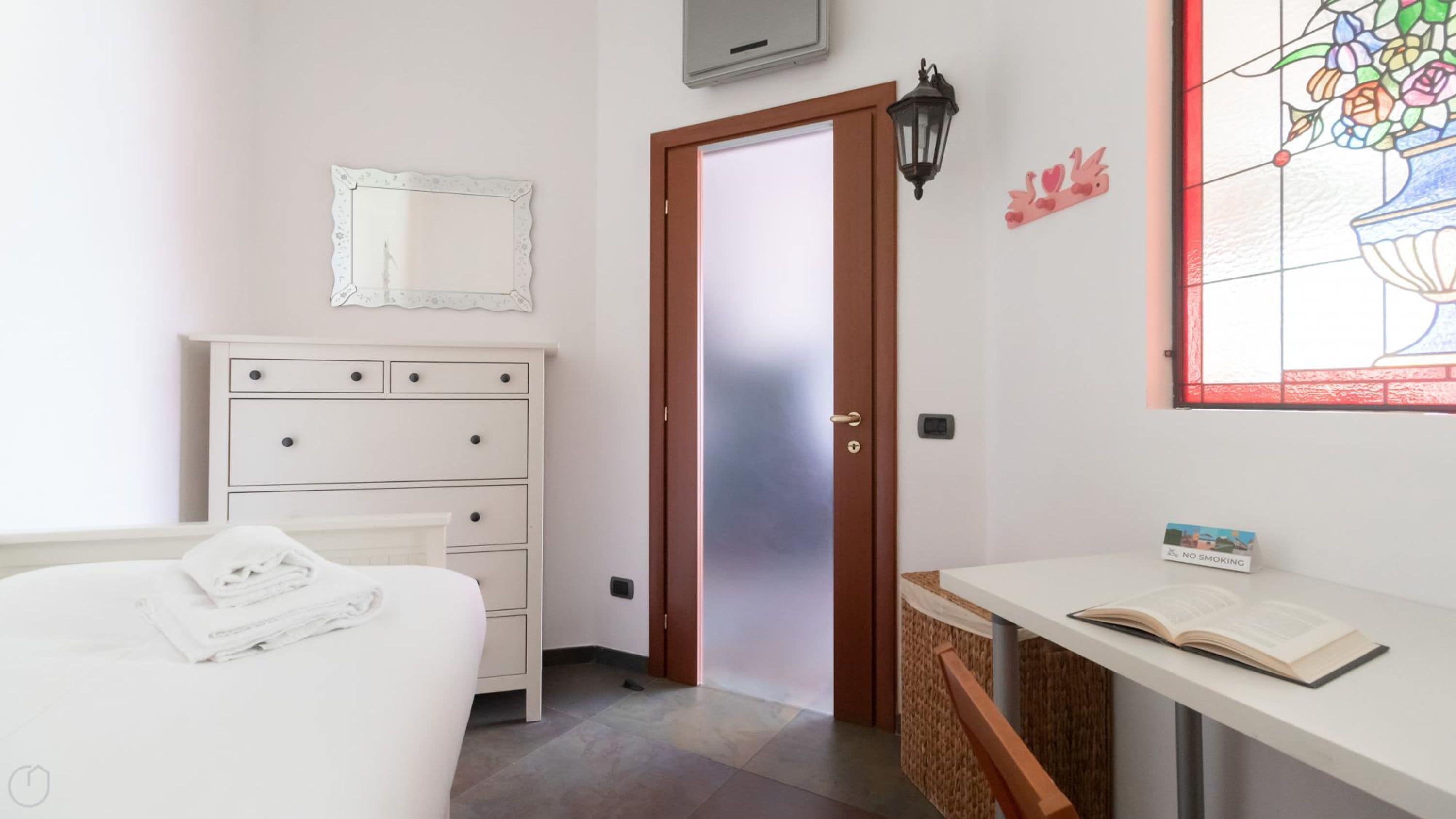 A bedroom with a bed, a dresser, a door, a wall-mounted light, a window with a stained glass design, and a bedside table with a book and a 'No Smoking' sign.