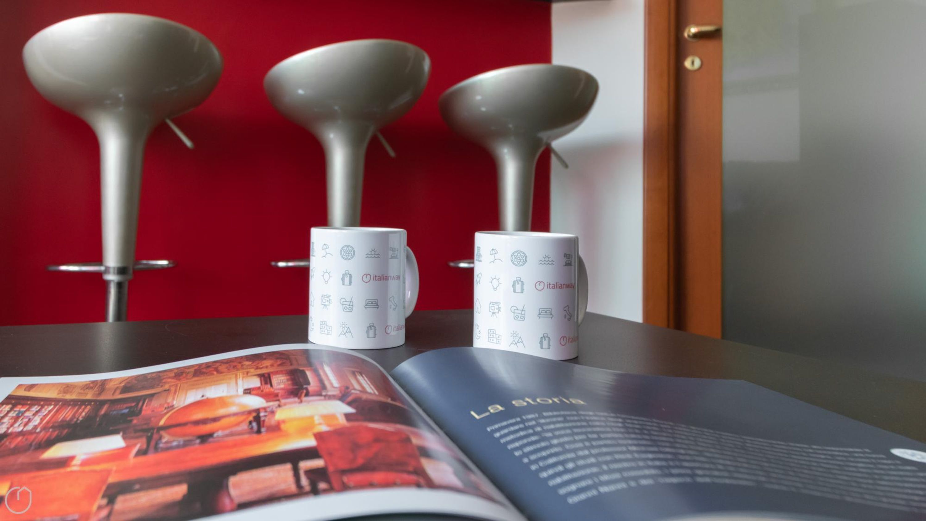 Three white mugs with Italian symbols on a table, a magazine with a red cover, and a door in the background.