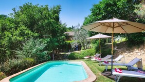 Une piscine entourée de chaises longues et de parasols, avec de la verdure tout autour et un bâtiment en arrière-plan.