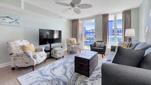 Una sala de estar con televisor de pantalla plana, sillones estampados, un sofá gris, una mesa de centro y vistas a edificios a través de ventanas amplias.
