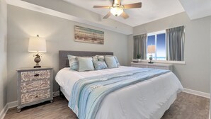 Un dormitorio con cama, mesita de noche, cómoda, ventilador de techo, ventana con cortinas y un cuadro en la pared.