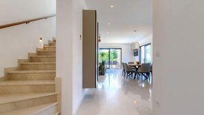Un intérieur moderne avec un escalier en marbre, un coin repas comprenant une table et des chaises, et une vue sur l’extérieur grâce à de grandes fenêtres.