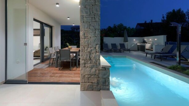 Un espace piscine extérieur moderne, agrémenté d’un pilier en pierre, d’un coin repas avec des chaises et une table, et offrant une vue sur le paysage environnant.