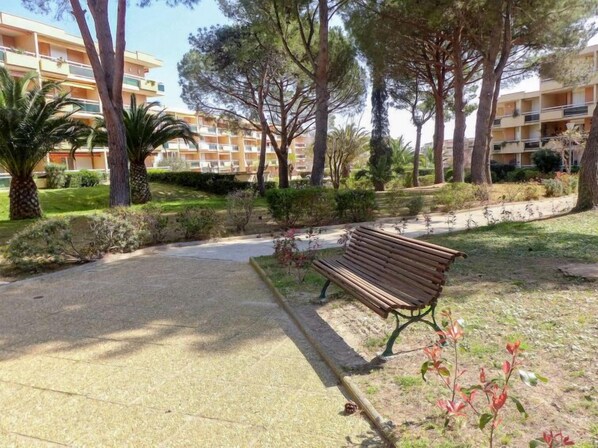 Un espace vert aménagé avec un banc, des arbres et un sentier menant à un bâtiment doté de balcons.