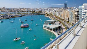 A marina with numerous boats, a modern building with a glass facade, and a cityscape in the background. - Superlative Penthouse With Valletta and Harbour Views (Sliema)