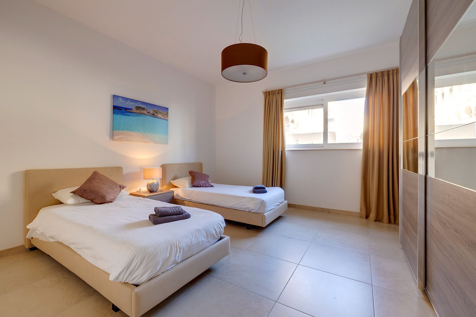 A hotel room with two beds, a painting, a lamp, and a view of the city through large windows.