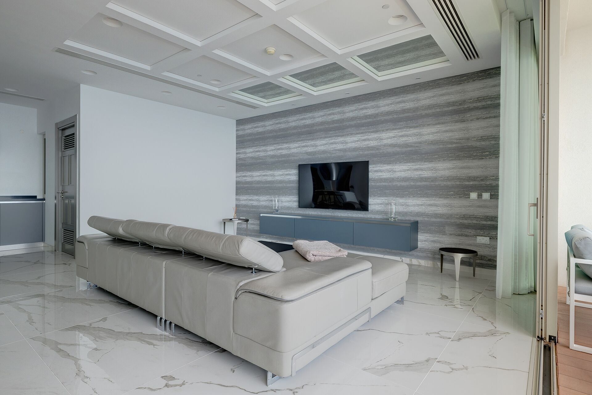 A modern living room with a grey sofa, a flat-screen TV mounted on a textured wall, and a minimalist design.