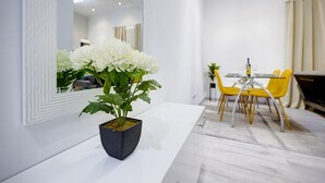 A modern living room with a white sofa, a dining table with yellow chairs, and a potted white flower plant on a white console table.