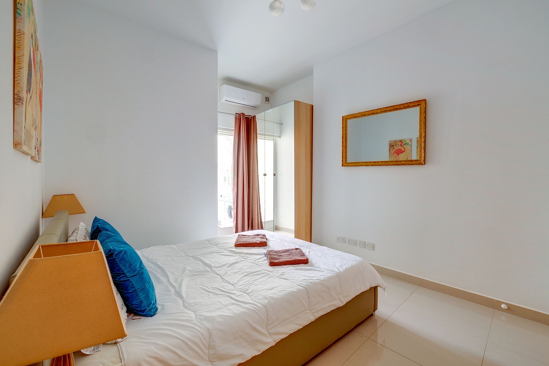 A bedroom with a bed, a lamp, a painting, and a mirror.