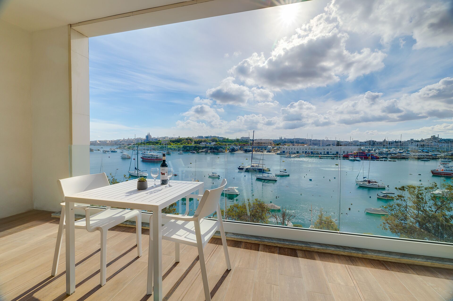 A balcony with a table and chairs, overlooking a marina with boats and a cityscape.