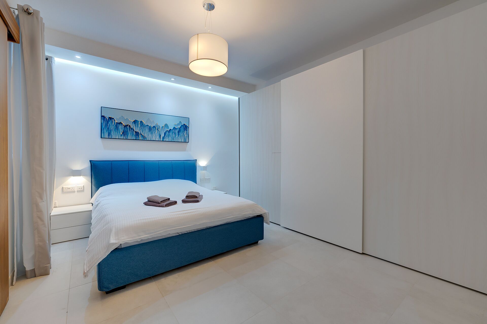 A modern bedroom with a large bed, a blue headboard, a painting on the wall, and a wardrobe.