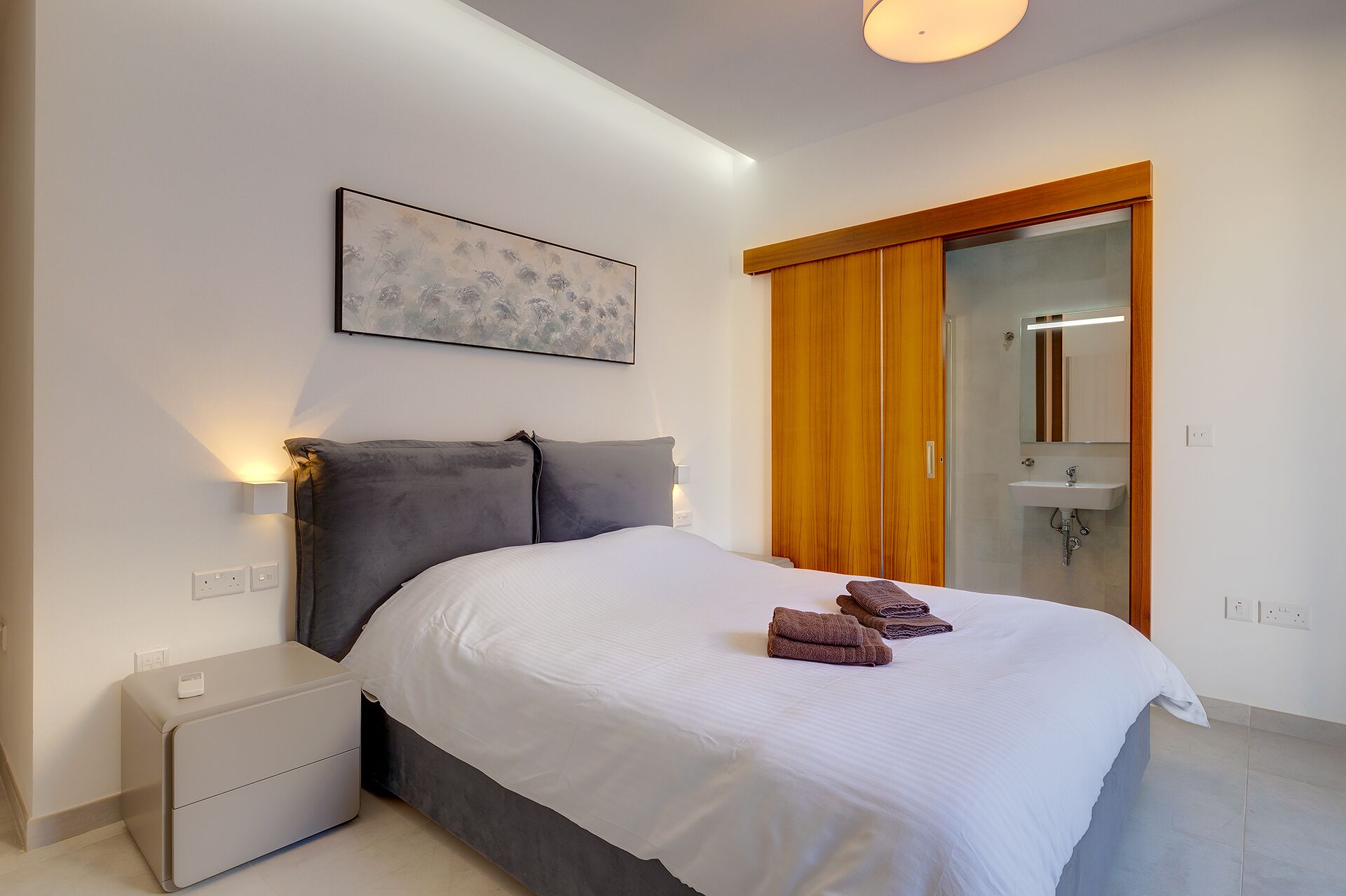 Een slaapkamer met een bed, nachtkastjes, een schilderij, een schuifdeur naar een badkamer en een plafondlamp.