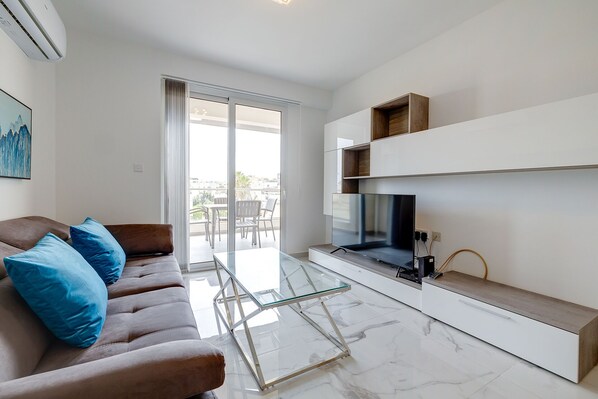 A modern living room with a glass coffee table, a brown sofa with blue cushions, a flat-screen TV on a wooden shelf, and a view of the city through large windows.