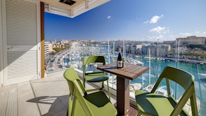 Een balkon met een tafel, stoelen en een fles wijn. Er is uitzicht op een jachthaven met boten en gebouwen.