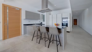 A modern kitchen with a central island, bar stools, and a view of the city through the window. - Luxury Apartment With Valletta and Harbour Views (Sliema)