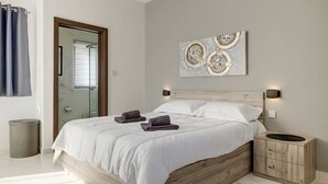 A bedroom with a wooden headboard, a bed with white bedding, two wall-mounted nightstands, a small wooden cabinet, and a bathroom visible through a glass door.