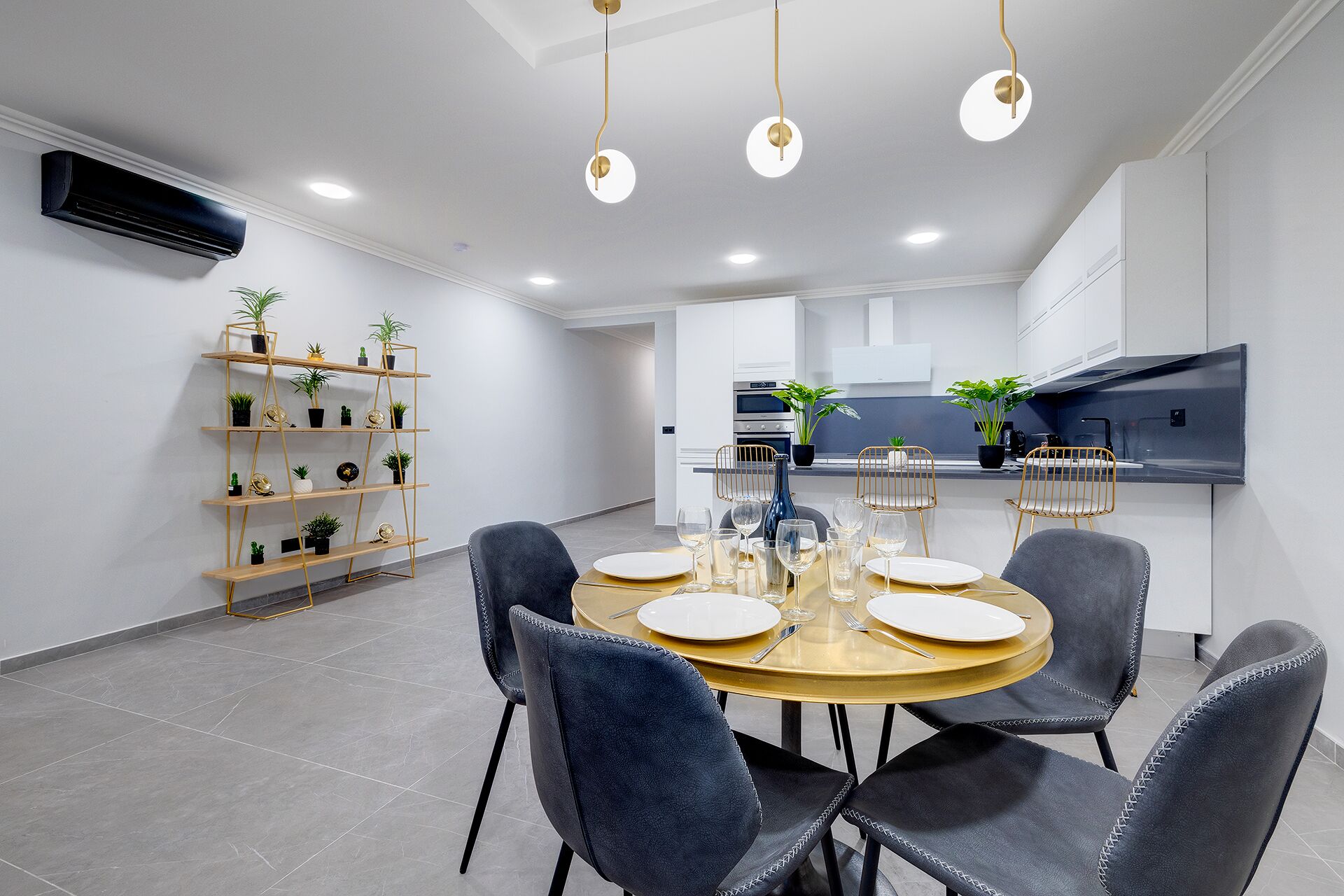 A modern dining area with a round table set for four, surrounded by dark chairs. There is a kitchen in the background with a large island and modern appliances.