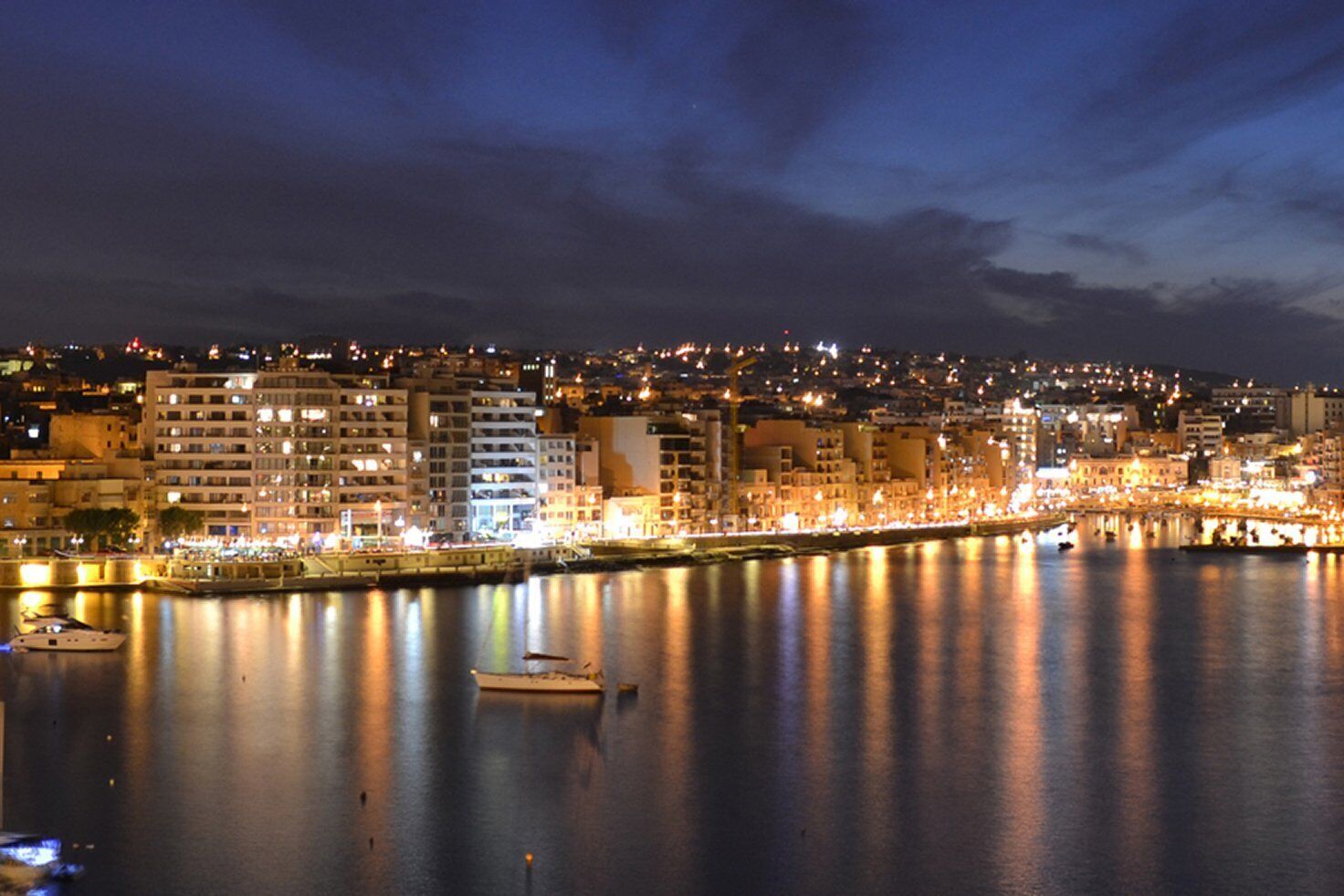 Paesaggio urbano notturno con edifici lungo una banchina, imbarcazioni sull'acqua e un fiume calmo che riflette le luci.