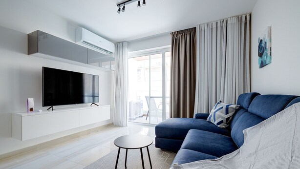 A modern living room with a flat-screen TV, a blue sofa, a small round table, and a large window with curtains. - Chic 2BR Apartment, Ideal Location (Ta' Xbiex)