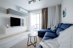 Ett modernt vardagsrum med en platt-TV, en blå soffa, ett litet runt bord och ett stort fönster med gardiner.